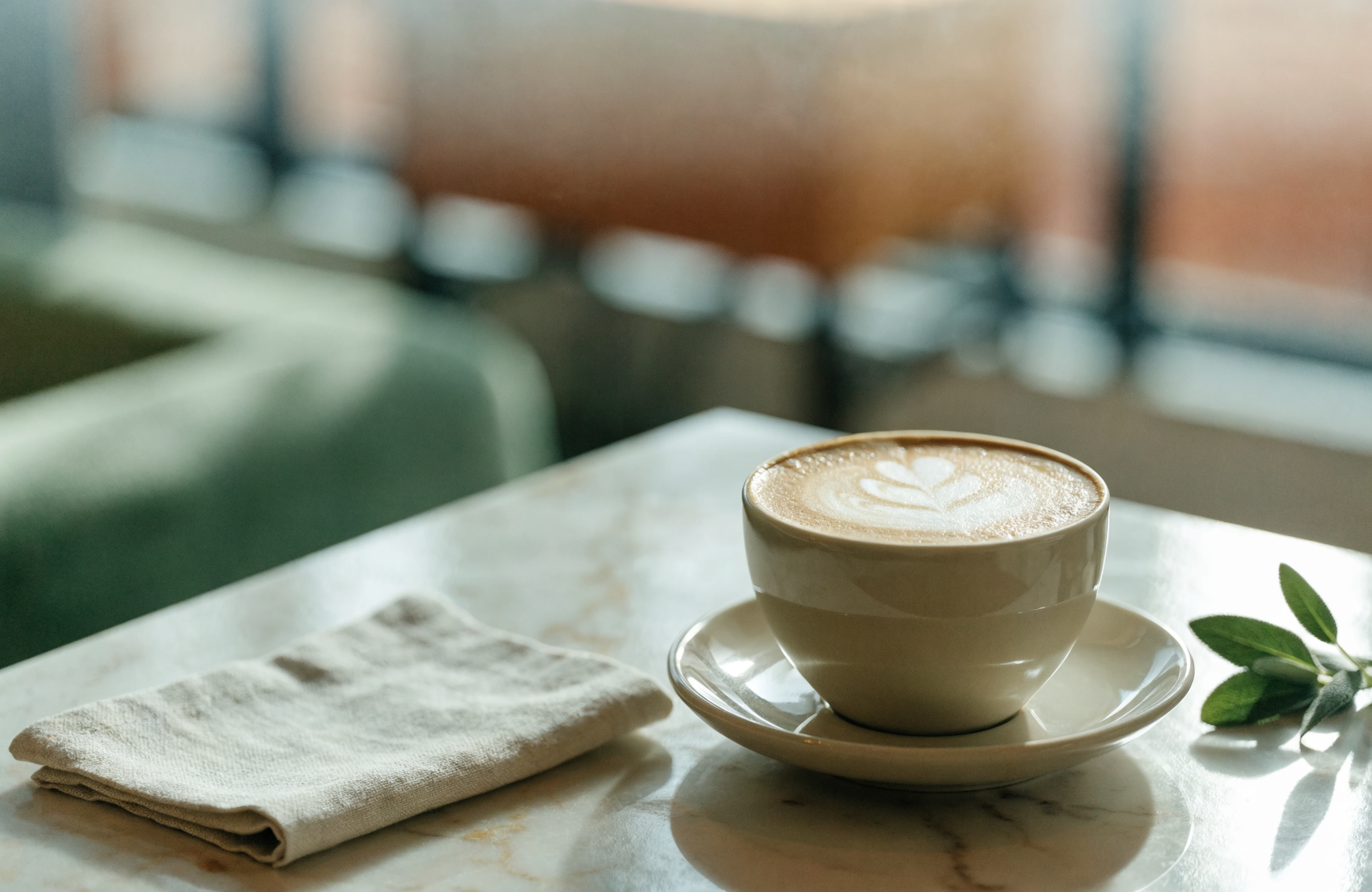 Cappuccino with rosetta latte art beside a folded linen napkin and a sprig of sage, on a marble table in morning light