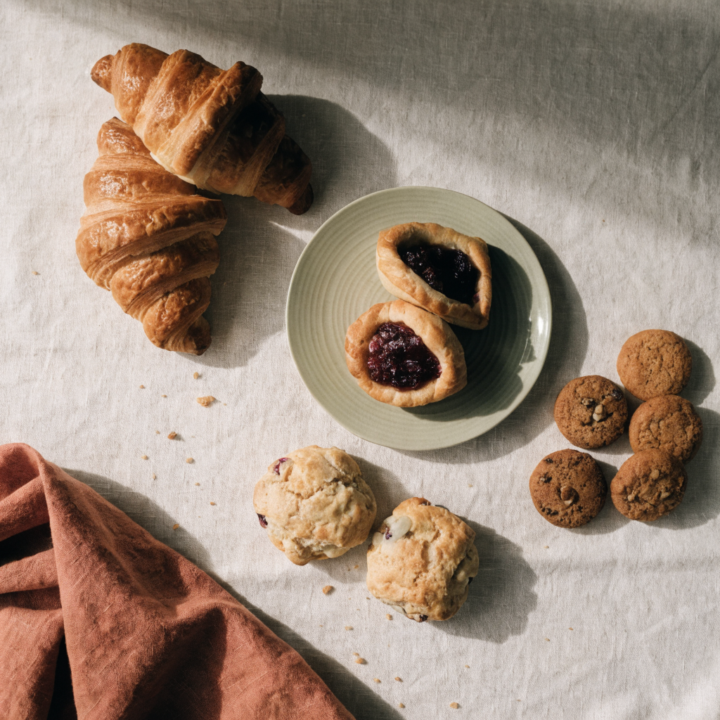 Overhead spread of croissants, danishes, scones, and cookies on linen with a sage plate and terracotta napkin