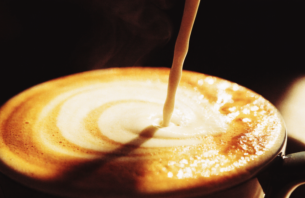Steamed milk pouring into espresso, latte art forming under warm gold light