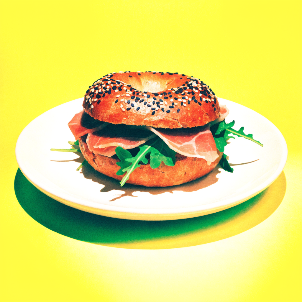 Seeded bagel sandwich with prosciutto and arugula on a yellow background